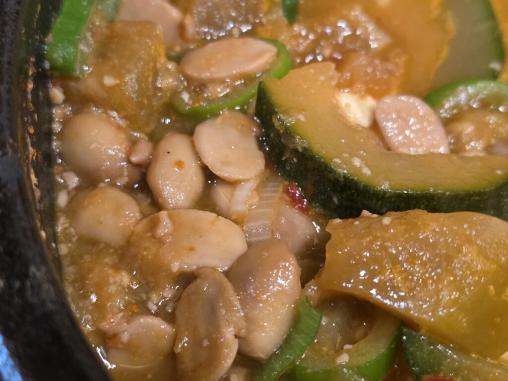 A close-up shot for a what is why how guide explained about Cheonggukjang, showing the thick texture of fermented whole soybeans, sliced zucchini, and rich broth in a stone pot.