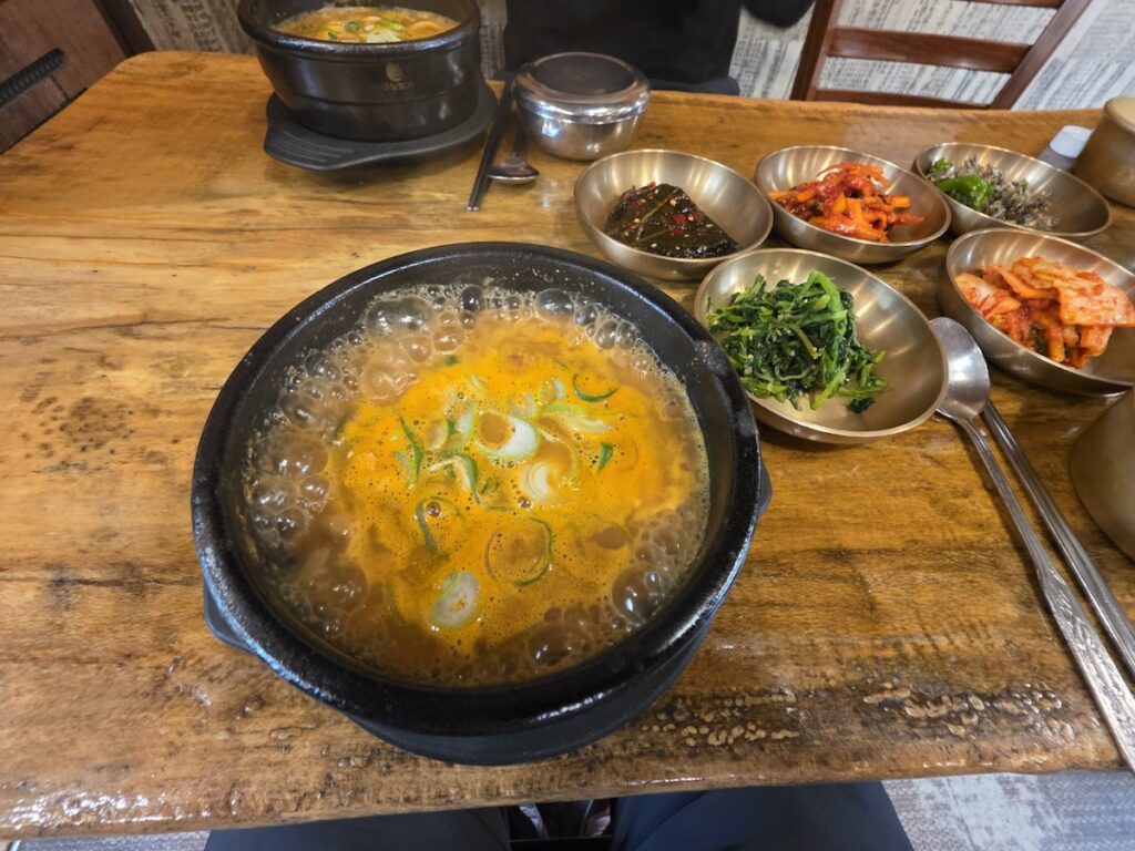 A full table setting featured in a what is why how guide explained, showcasing a bubbling Cheonggukjang pot, brass bowls with side dishes like kimchi and seasoned greens, and a traditional wooden table.