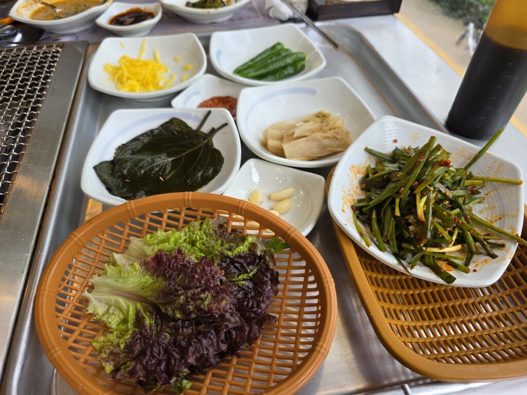 A variety of Korean side dishes (banchan) including raw ginger, chives, and perilla leaves served with eel.