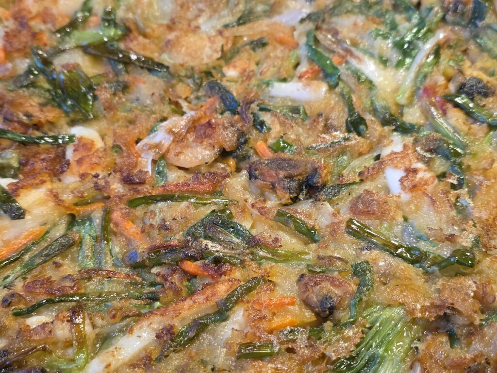 A close-up, high-angle shot showing the crispy texture of a Myeongdong Seafood Pancake, featuring visible green onions, sliced carrots, and pieces of seafood embedded in the golden-brown batter.