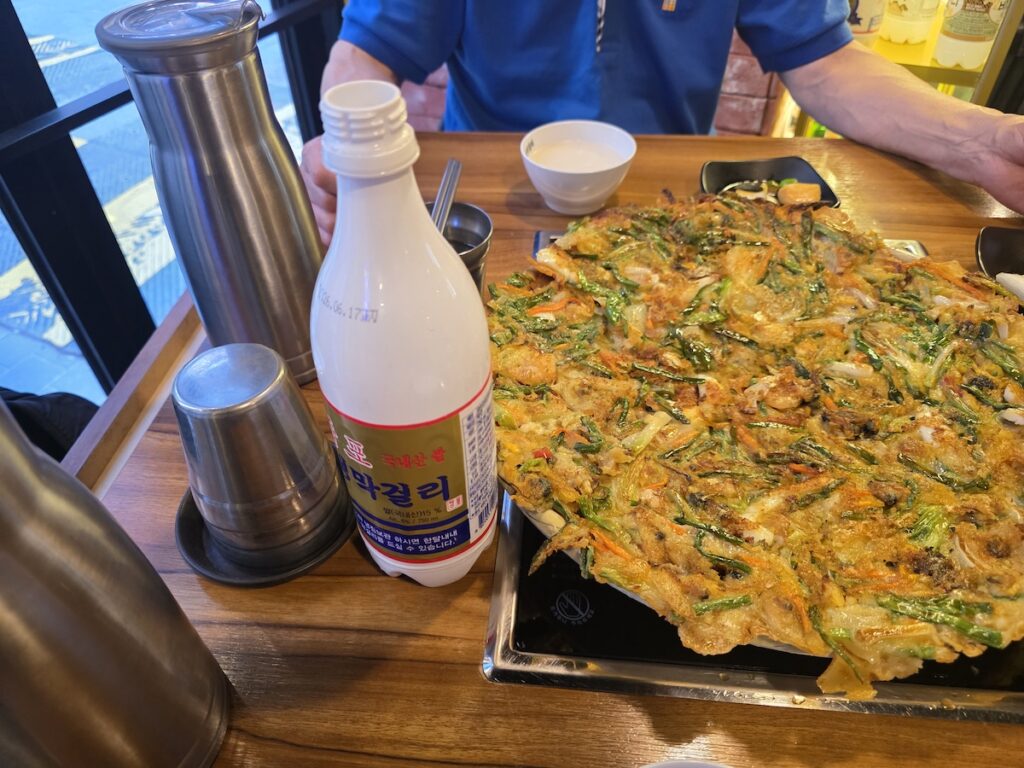 A view of a wooden dining table with a bottle of makgeolli in the foreground and a person sitting in the background, set in a restaurant where Myeongdong Seafood Pancake is served.