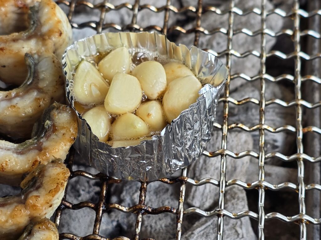Fresh garlic cloves cooking in a small oil-filled foil cup on a grill next to Korean freshwater eel.