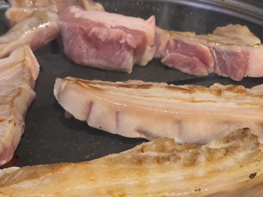 Slices of pork belly browning on a charcoal grill, showing the searing process and grill marks.