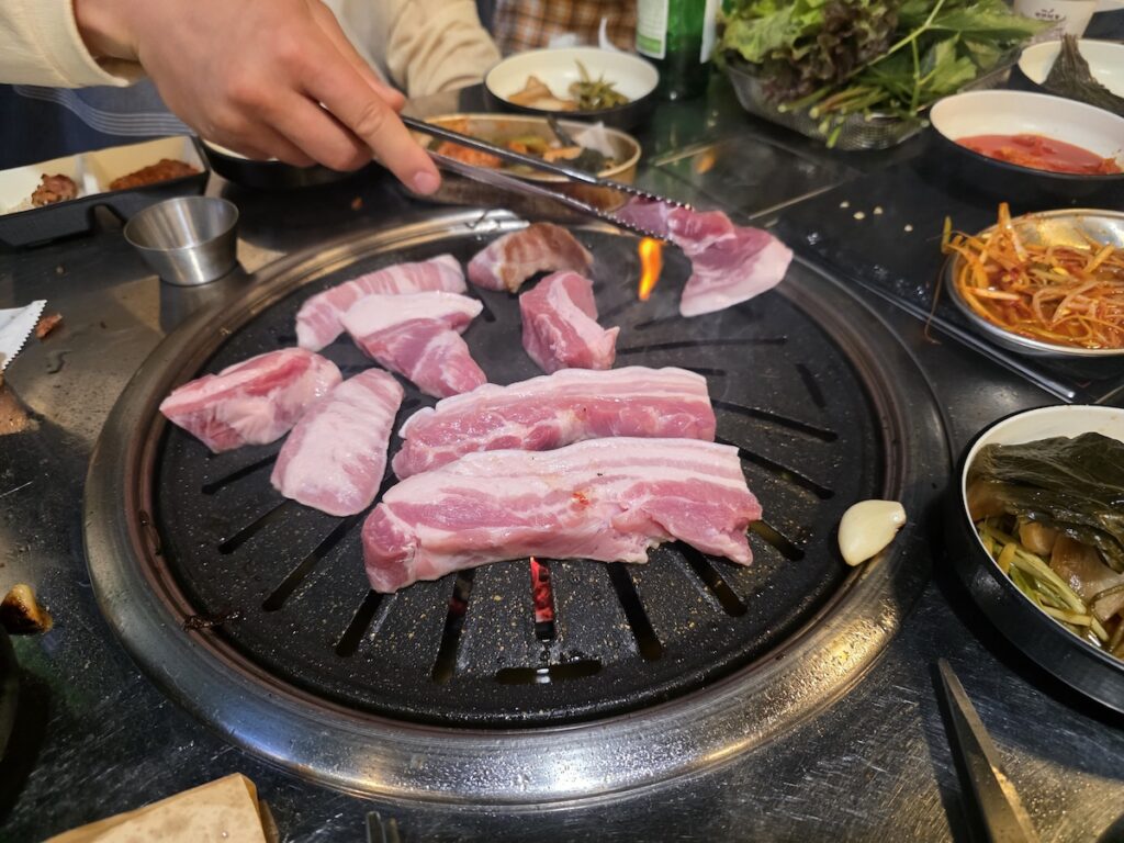 A person using tongs to place thick slices of raw pork belly (Samgyeopsal) on a round grill at a Korean BBQ restaurant in Seoul.