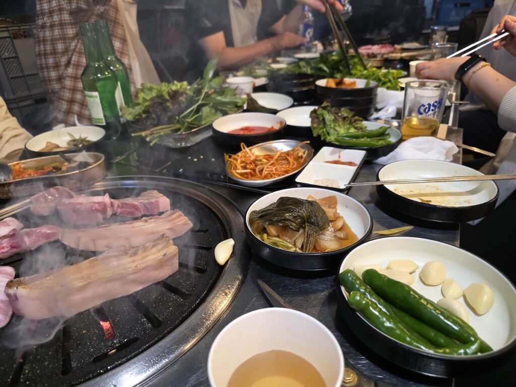 A full Korean BBQ table setting with various side dishes (Banchan), fresh lettuce, garlic, green peppers, and bottles of Soju and Beer.