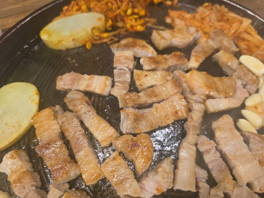A person using metal tongs to arrange raw samgyeopsal (pork belly) on a hot round grill surrounded by Korean side dishes.