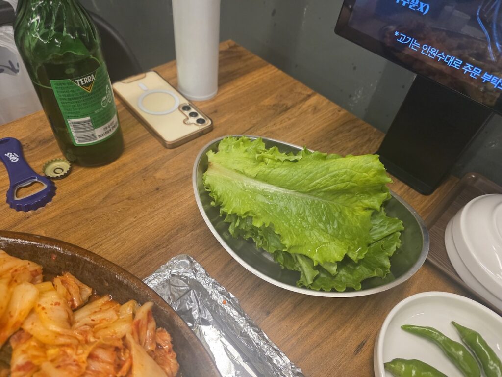 A crowded Korean BBQ table with green soju bottles, garlic, lettuce, scallion salad, and smoke rising from the central grill.
