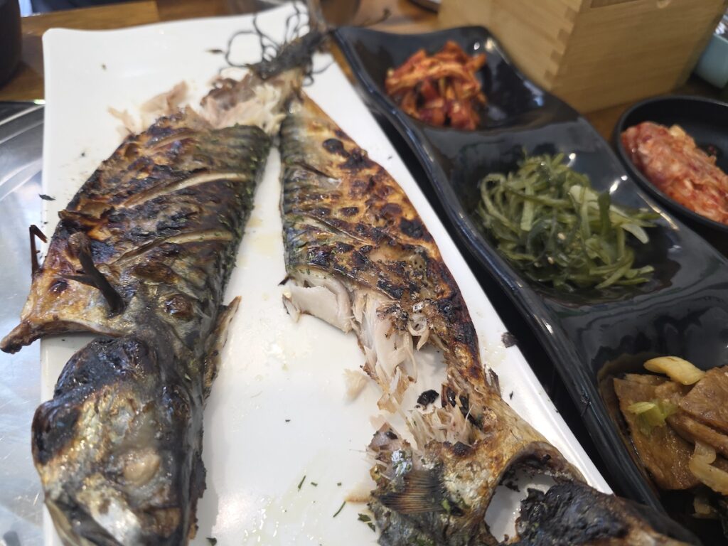 Korean grilled mackerel with side dishes on a restaurant table, showing a partially eaten fish with bones, crispy skin, and small banchan plates