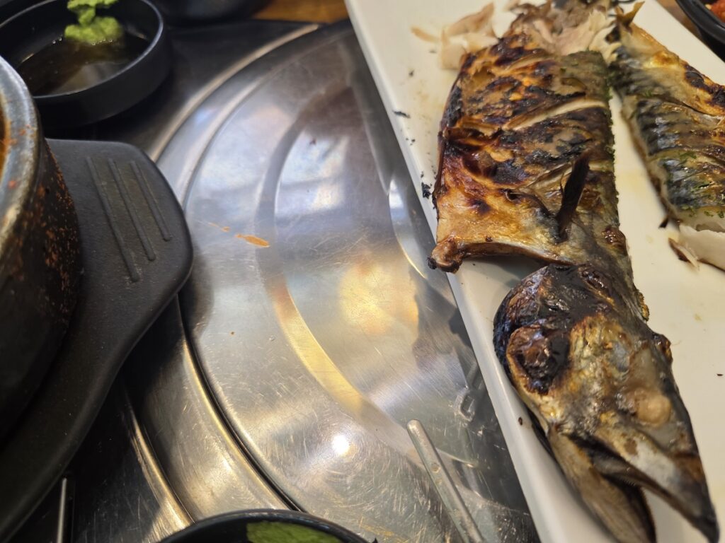 Korean grilled mackerel served whole on a white plate at a restaurant table, showing the head, skin, and bones that diners remove while eating
