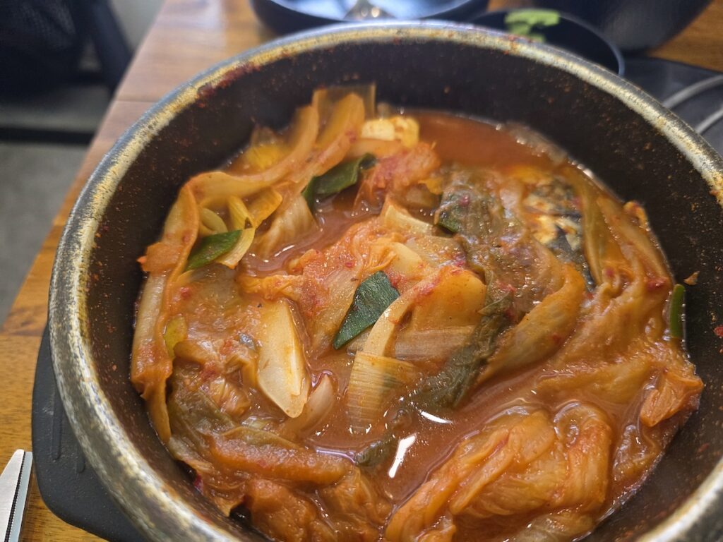 A bowl of Korean mackerel kimchi stew with bone-in fish, kimchi, onions, and spicy red broth served at a casual restaurant table