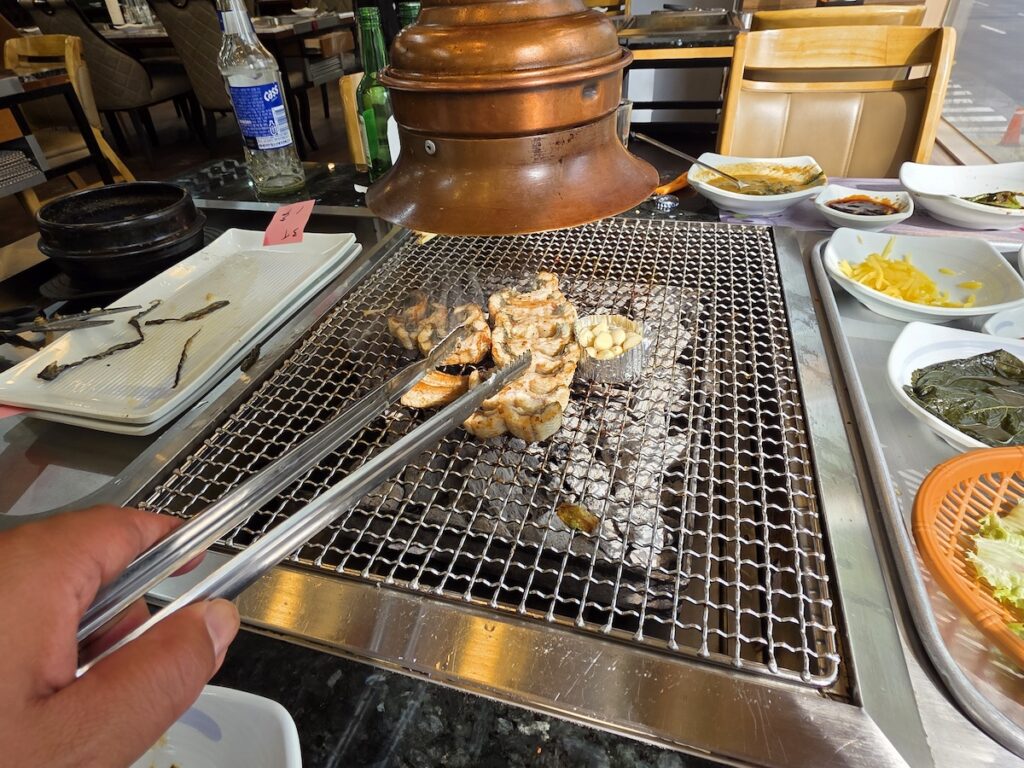 A wide shot of a Korean eel BBQ restaurant table with tongs flipping the eel over charcoal and various side dishes.