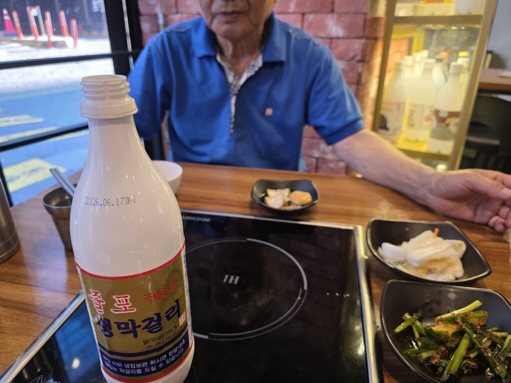A brightly lit glass display shelf stocked with various bottles of Korean rice wine, showcasing a wide selection of drinks to enjoy alongside a Myeongdong Seafood Pancake.