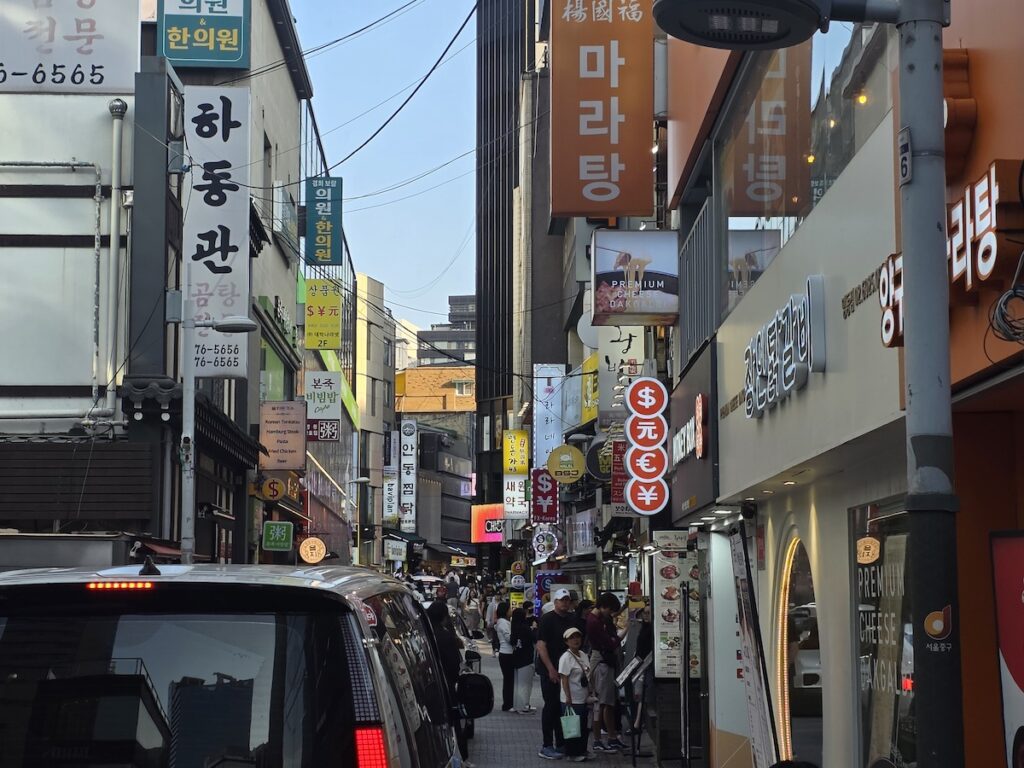 A bustling narrow street in Myeongdong, Seoul, crowded with pedestrians and lined with various restaurant signs, shops, and currency exchange symbols, leading toward popular dining areas known for Myeongdong Seafood Pancake.