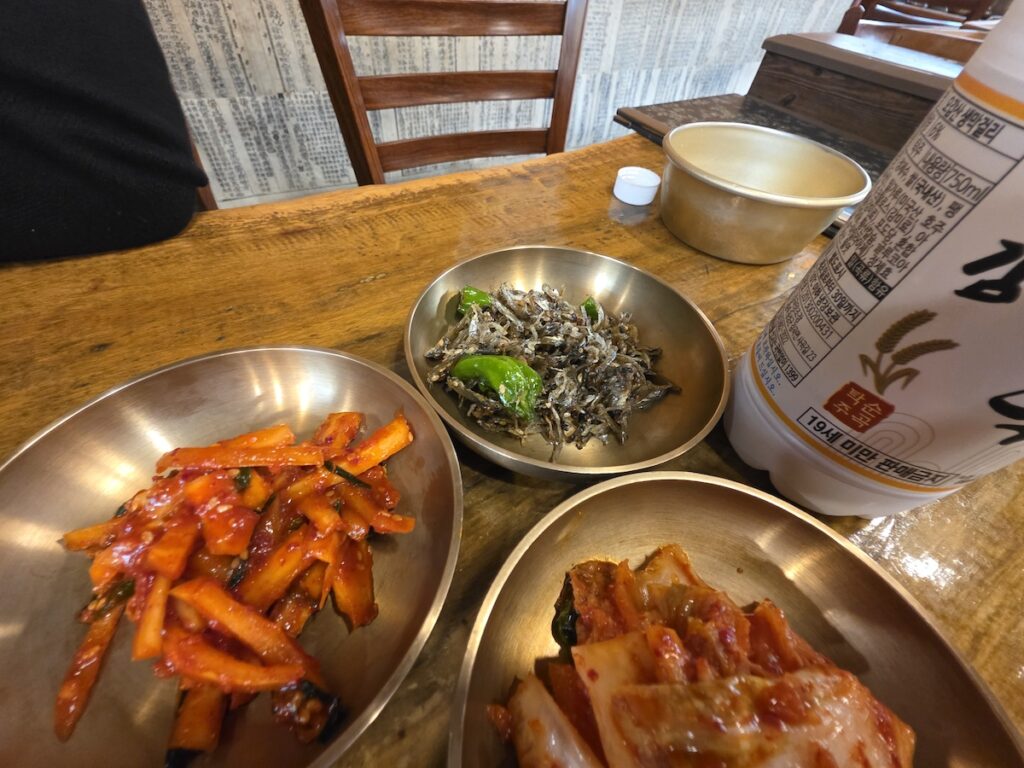 A variety of Korean side dishes including stir-fried dried anchovies (myeolchi-bokkeum) next to a bottle of Makgeolli rice wine.