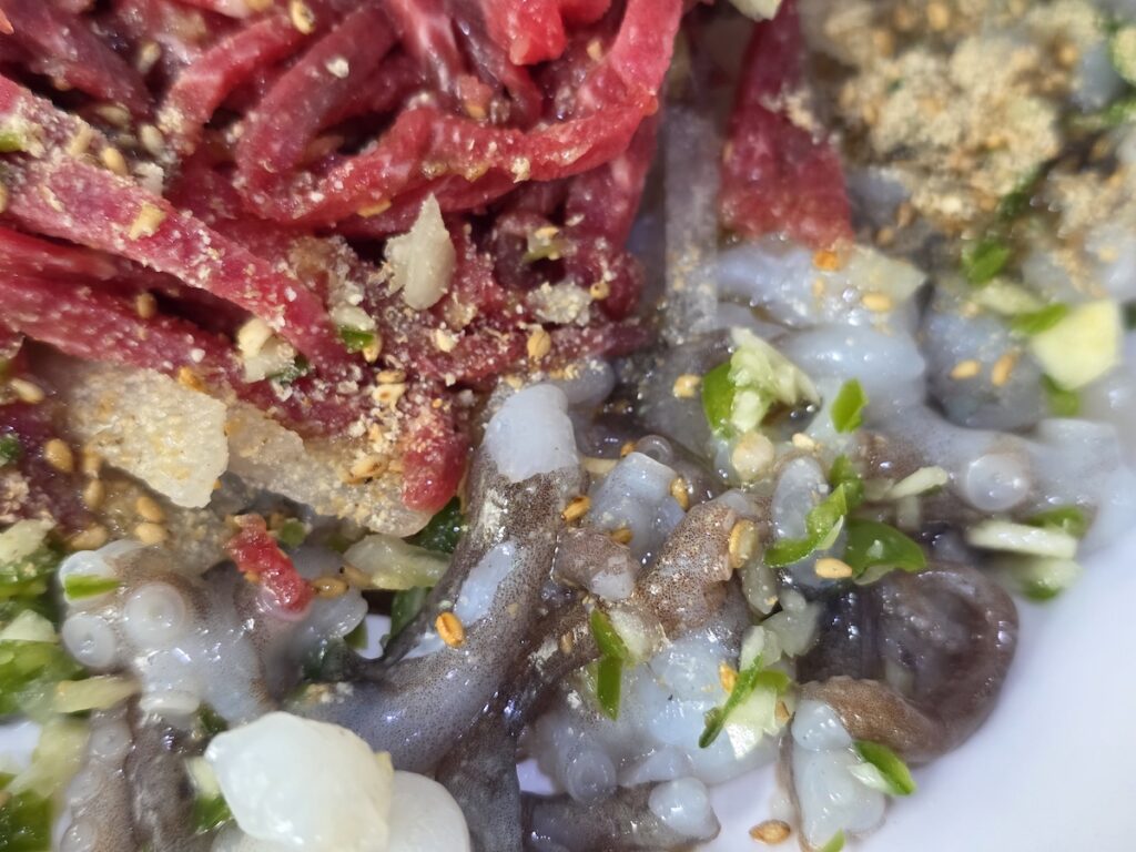 Close-up of chopped live octopus and seasoned raw beef, showing suckers, sesame seeds and green chili